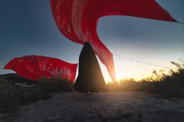 تحت راية العباس عليه السلام، وقفت السيدة زينب، لم تكن الراية قطعة قماش، بل كانت وصيّة أخٍ مضى دون أن يغيب، وعهداً مكتوباً بكفه المبتورة على صفحة الريح: أن تظلّ زينب مصونة، ولو بقي من العباس ظلّ راية فقط